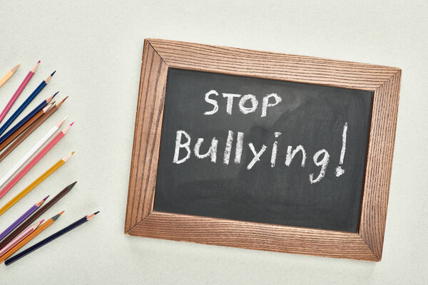 top view of chalkboard in wooden frame with stop bullying lettering near colored pencils on grey background