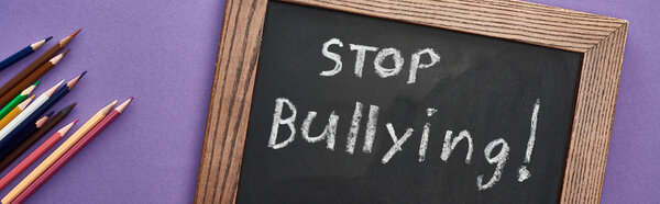 panoramic shot of chalkboard in wooden frame with stop bullying lettering near colored pencils on purple background