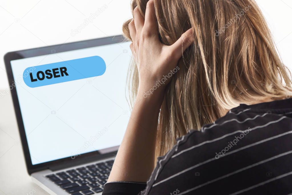 offended blonde girl as victim of cyberbullying sitting near laptop with loser message on screen