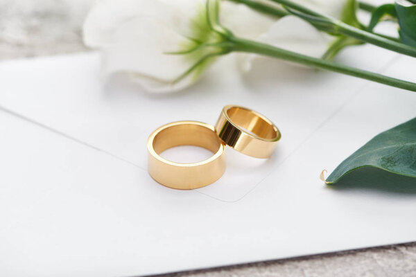 golden wedding rings on white envelope near white flowers