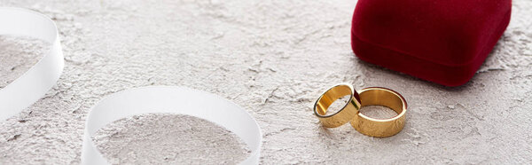 panoramic shot of golden rings near gift box and white ribbon on textured surface