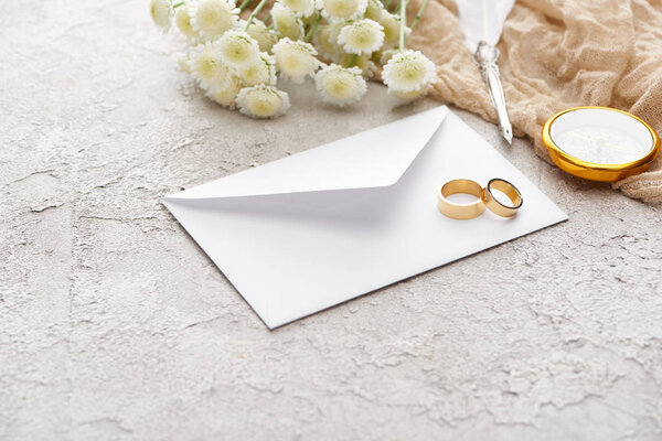 golden rings on white envelope near chrysanthemums, beige sackcloth, quill pen and compass on textured surface 