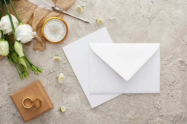 top view of white envelope, compass, wedding rings on gift box on grey textured surface