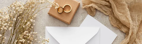 在纹理表面上的礼品盒上 白色信封和卡片的全景拍摄 靠近鲜花 米色麻布和结婚戒指 — 图库照片