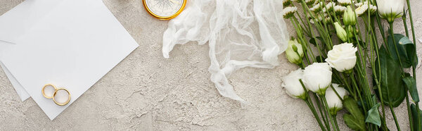 panoramic shot of wedding rings on blank invitation card, white cheesecloth and flowers on textured surface
