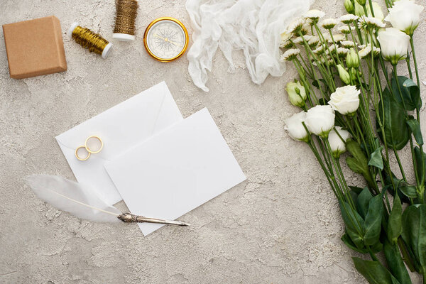 top view of empty card near wedding rings, quill pen, compass, cheesecloth and flowers on textured surface