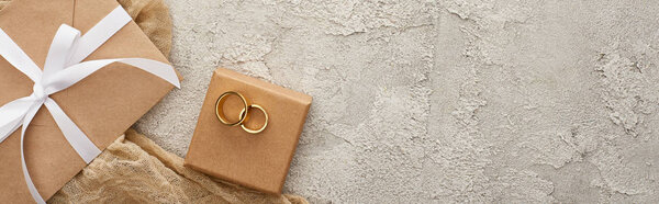 panoramic shot of wedding rings on gift box near beige envelope with white ribbon near sackcloth on textured surface 