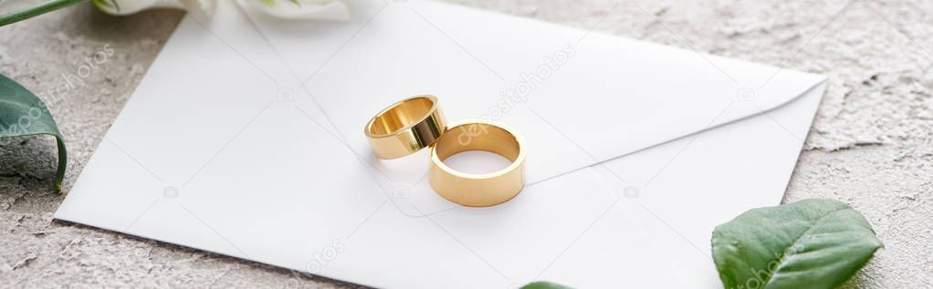panoramic shot of golden rings on white envelope near roses on grey textured