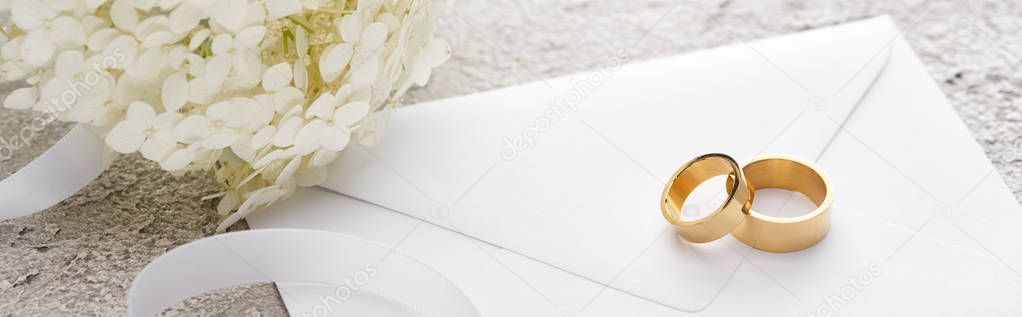 panoramic shot of golden rings on envelope near white ribbon and Hortense flower on grey textured surface