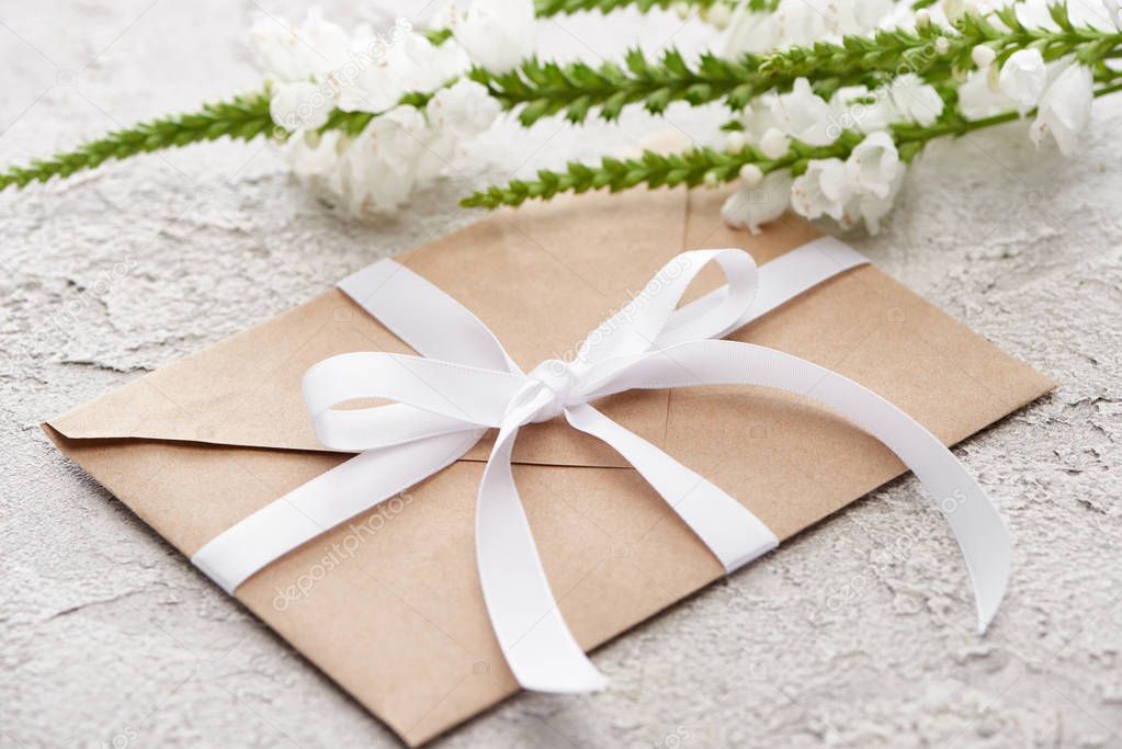 Beige envelope with white ribbon near flowers on grey textured surface