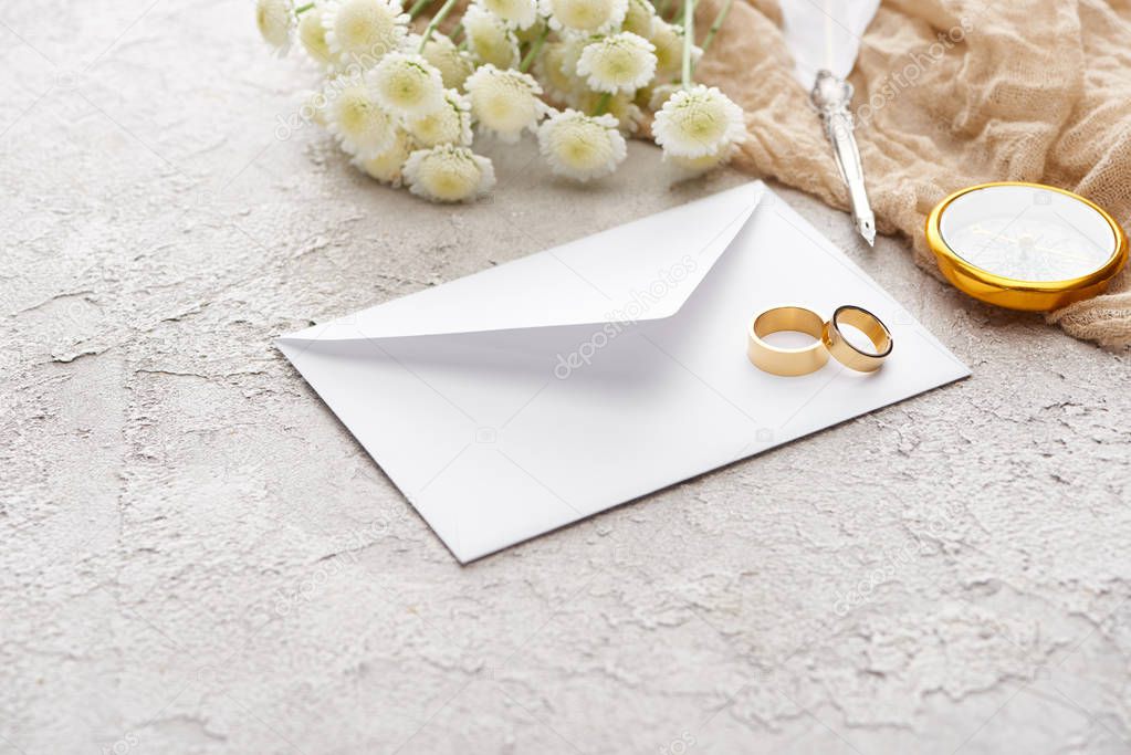 golden rings on white envelope near chrysanthemums, beige sackcloth, quill pen and compass on textured surface 