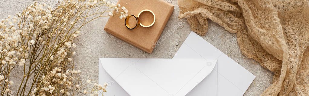 panoramic shot of white envelope and card near flowers, beige sackcloth and wedding rings on gift box on textured surface