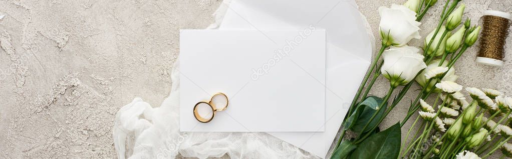 panoramic shot of golden rings on blank card near envelope on white cheesecloth and eustoma flowers on textured surface