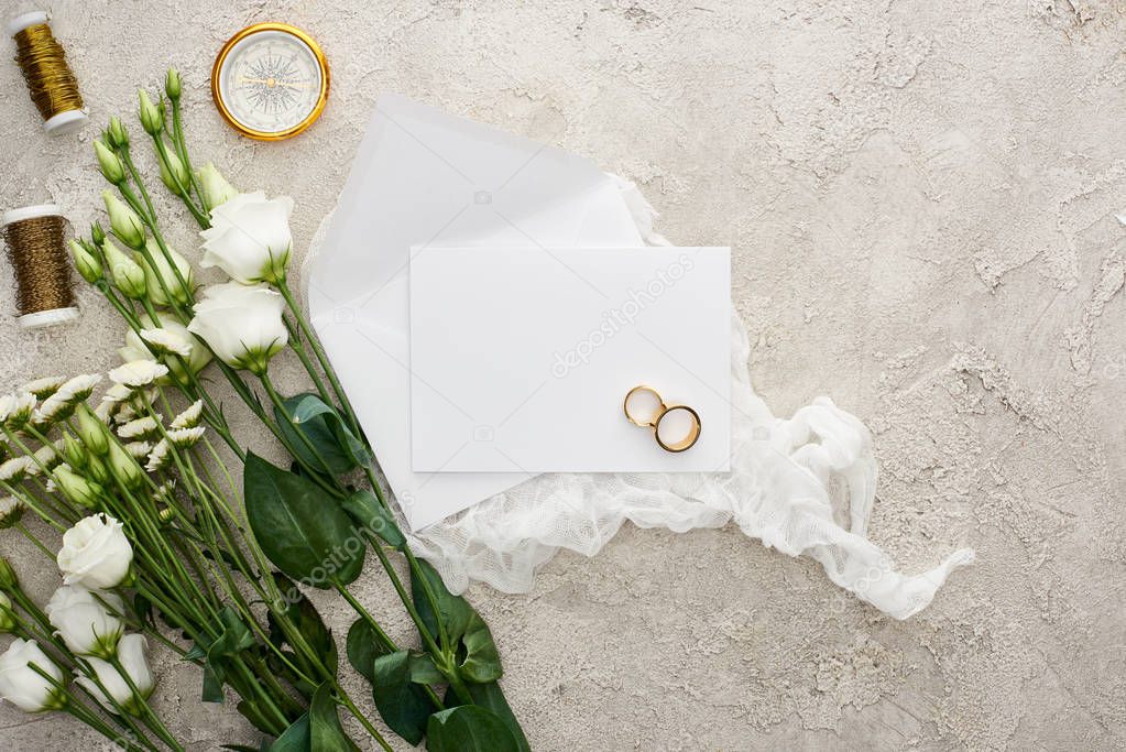 top view of eustoma flowers near wedding rings on empty card, white cheesecloth, golden compass and bobbins on grey textured surface 