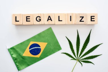 top view of green cannabis leaf and legalize lettering on wooden cubes near flag of Brazil on white background clipart