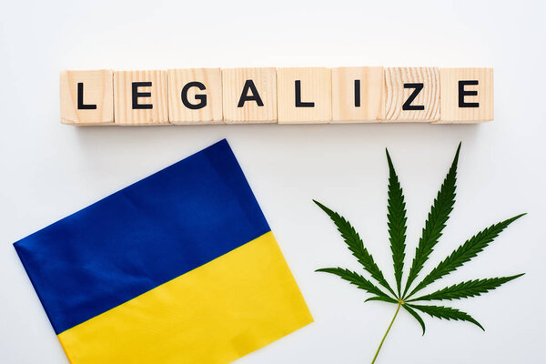 top view of green cannabis leaf and legalize lettering on wooden cubes near flag of Ukraine on white background