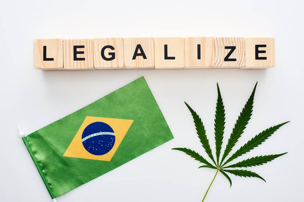 top view of green cannabis leaf and legalize lettering on wooden cubes near flag of Brazil on white background
