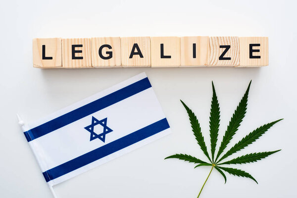 top view of green cannabis leaf and legalize lettering on wooden cubes near flag of Israel on white background