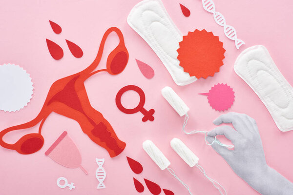 обрезанный вид белой руки, держащей тампон на розовом фоне с гигиеническими салфетками, разрезанной бумагой женских репродуктивных внутренних органов и капель крови
