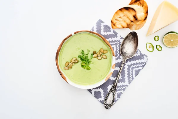 おいしいクリーミーな緑色の野菜スープのトップビューは 白に分離されたグリルクルトンとチーズとスプーンの近くにナプキンで提供 — ストック写真