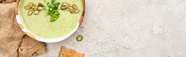 top view of tasty green creamy soup with crackers on textured grey background with rustic cloth, panoramic shot
