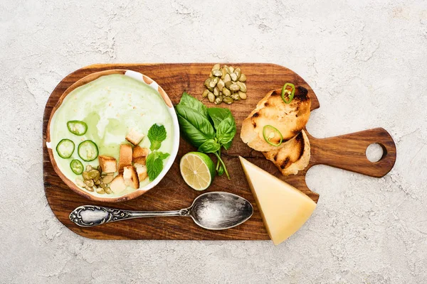 Top view of green vegetable creamy soup on wooden chopping board near cheese, croutons and spoon on textured surface — Stock Image Top View Green Vegetable Creamy Soup Wooden Chopping Board Cheese Stock Image