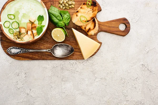 Top view of green vegetable creamy soup on wooden chopping board near cheese and spoon on textured surface — Stock Image Top View Green Vegetable Creamy Soup Wooden Chopping Board Cheese Stock Picture