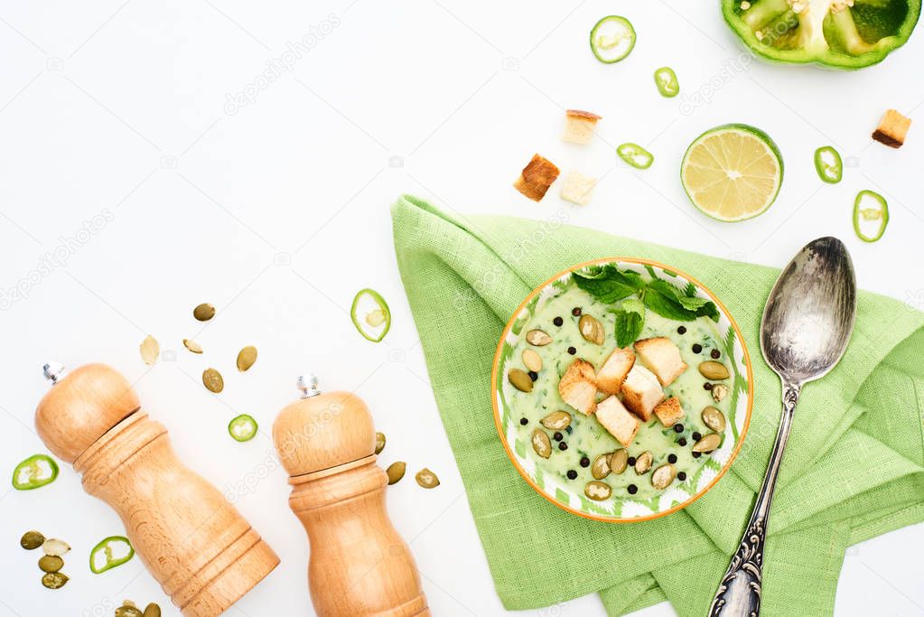 top view of delicious creamy green vegetable soup served with salt and pepper mills, spoon and napkin isolated on white