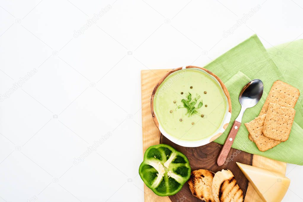 top view of delicious creamy green vegetable soup served on wooden cutting board with croutons and cheese isolated on white