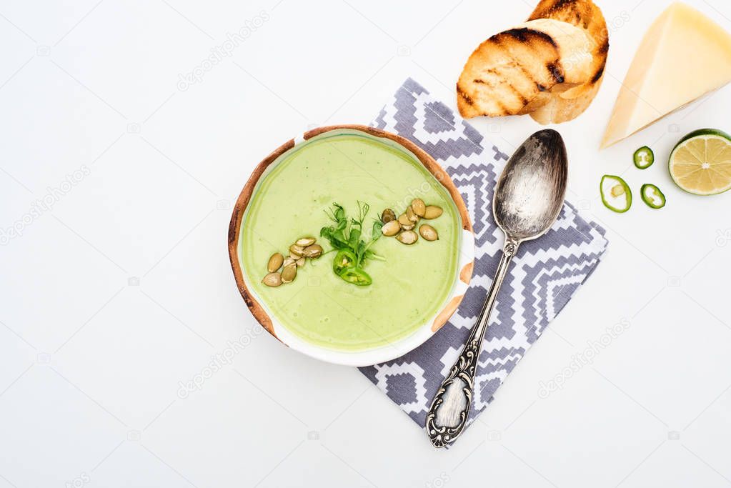 top view of delicious creamy green vegetable soup served on napkin near spoon with grilled croutons and cheese isolated on white