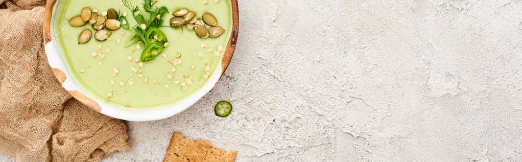 top view of tasty green creamy soup with crackers on textured grey background with rustic cloth, panoramic shot