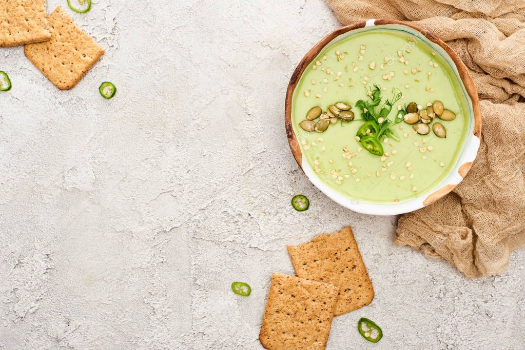 top view of tasty green creamy soup with crackers on textured grey background with rustic cloth