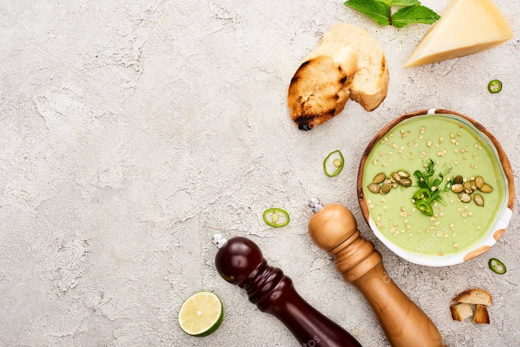 top view of tasty green creamy soup with croutons, cheese and salt and pepper mills on textured grey background
