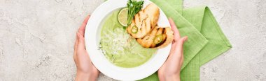 cropped view of woman holding plate with delicious creamy green vegetable soup with croutons near green napkin, panoramic shot clipart