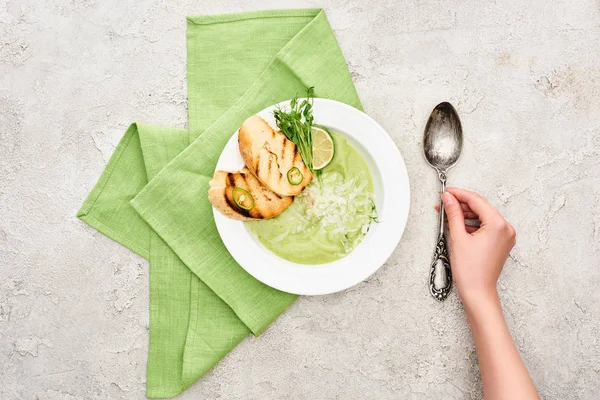 クルトンと一緒に出されるおいしいクリーミーな緑の野菜スープの近くにスプーンを入れる女性の部分的なビュー — ストック写真