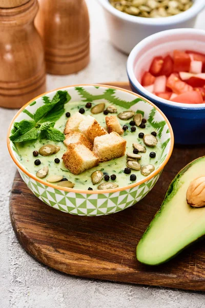 Bowl of delicious green vegetable creamy soup with croutons, black pepper and pumpkin seeds near avocado — Stock Image Bowl Delicious Green Vegetable Creamy Soup Croutons Black Pepper Pumpkin Stock Photo