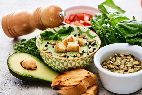 Bowl of delicious green vegetable creamy soup with croutons, black pepper and pumpkin seeds near fresh ingredients — Stock Image Bowl Delicious Green Vegetable Creamy Soup Croutons Black Pepper Pumpkin Stock Picture