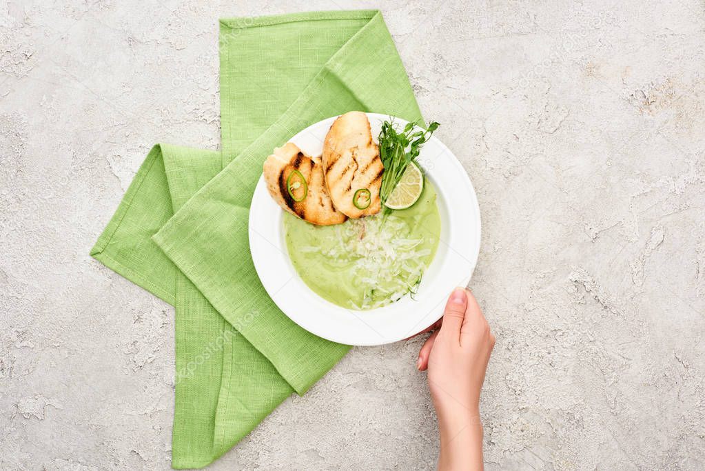 partial view of woman holding plate with delicious creamy green vegetable soup with croutons