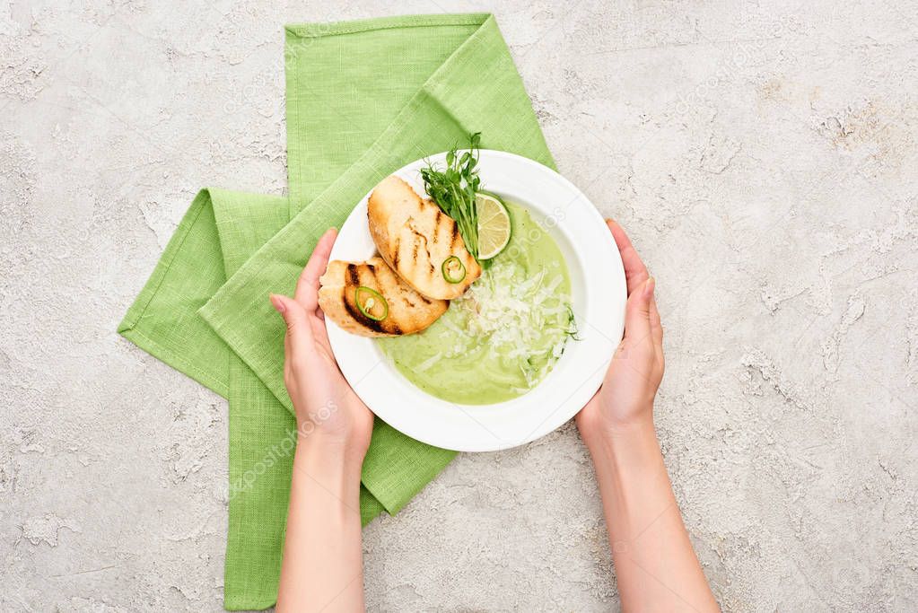 cropped view of woman holding plate with delicious creamy green vegetable soup with croutons near green napkin