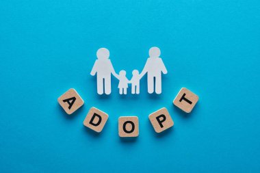 top view of paper cut gay family holding hands on blue background with adopt word on wooden cubes clipart