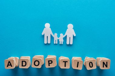 top view of paper cut gay family holding hands on blue background with adoption lettering on wooden cubes clipart