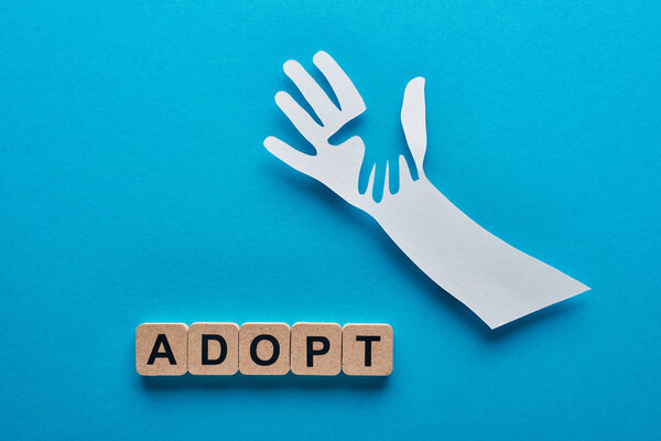 top view of paper cut parent and child hands near wooden cubes with adopt lettering on blue background