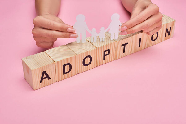 cropped view of woman holding paper cut family on wooden cubes with adoption lettering on pink background