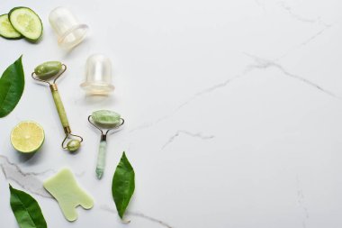 top view of nephrite massage rollers, vacuum jars, spatula, leaves, cucumber and lime on marble surface clipart