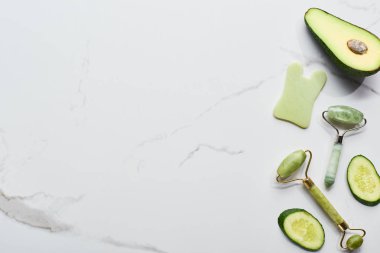 top view of facial spatula and rollers near avocado and cucumber on marble surface clipart