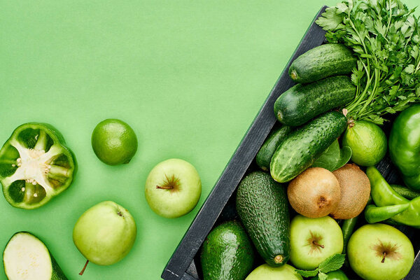 top view of apples, avocados, cucumbers, peppers, kiwi, greenery in wooden box