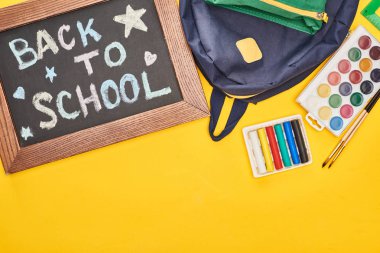 Chalkboard in wooden frame with back to school inscription near blue schoolbag and watercolor paints set on yellow background clipart