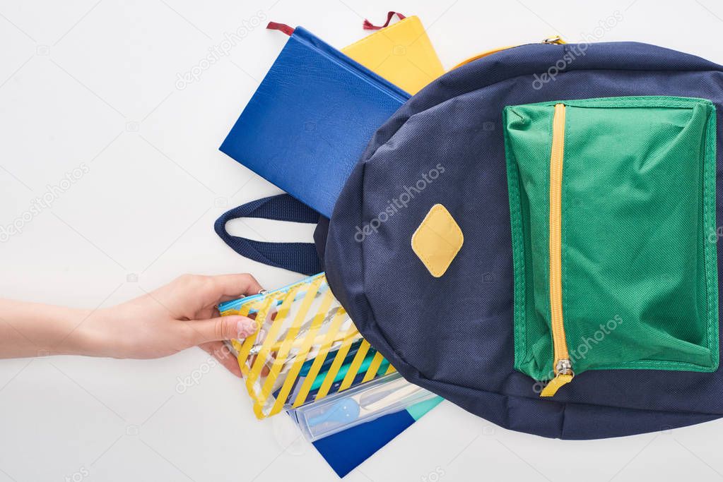 Cropped view of schoolgirl taking yellow pencil case from blue schoolbag with notepads isolated on white