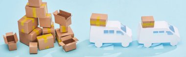 panoramic shot of pile of cardboard boxes near mini trucks on blue background clipart