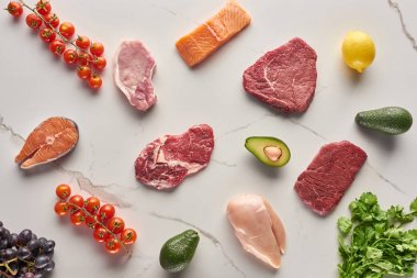 Gri mermer yüzeyde maydanoz, üzüm, kiraz domates, avokado ve limon yakınında çeşitli et, kümes hayvanları ve balık üst görünümü